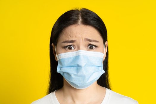 Scared of pandemic mature asian woman wearing medical face mask coronavirus or monkeypox prevention. Charming middle age woman in white t-shirt and medical mask on yellow background
