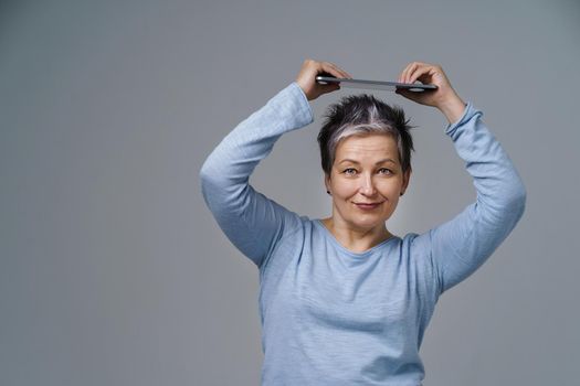 Playful grey haired mature woman holding digital tablet above head working or checking on social media. Pretty woman in 50s in blue blouse isolated on white background. Older people and technologies