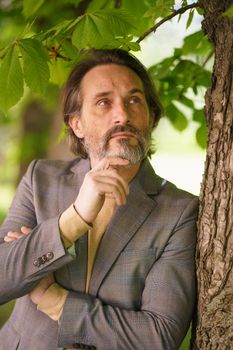 Mature handsome man with grey beard looking up leaning on tree wearing casual grey jacket. Life after 40 years concept, problems and depression. Middle age crisis