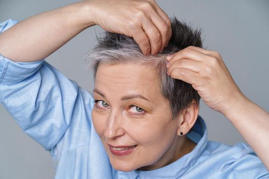 Pretty woman in 50s checking her hair or skull skin in mirror. Beautiful grey haired woman dealing with dandruff problem checking in mirror. Healthy hair and lifestyle concept