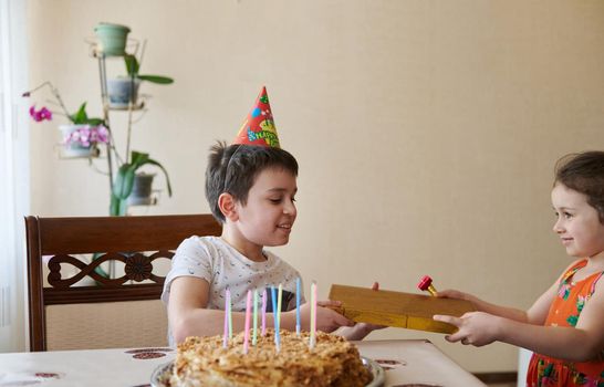 Charming Caucasian boy receives a gift from his younger sister for his 10th birthday, sits at the table with a birthday cake and candles. Birthday, anniversary and event celebration concept