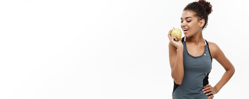 Healthy and Fitness concept - Beautiful American African lady in grey fitness clothes eating green apple. Isolated on white background