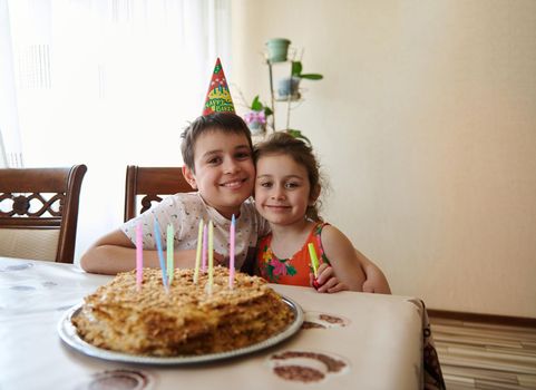 Portrait of two adorable cute children, brother and sister, sitting at the table with a birthday cake decorated with long colorful candles. Anniversary and birthday party concept