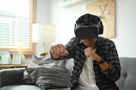 Joyful asian man wearing virtual reality headset playing simulation boxing game on weekend at home.