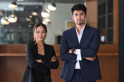 Two successful businesspeople in formal wear standing with crossed arms in modern office.