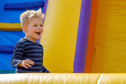 Kid jump in inflatable bounce castle on kindergarten birthday party. Activity and play center for young child. Little smiling boy playing outdoors in summer.