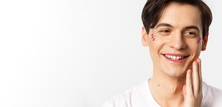 People, lgbtq and beauty concept. Headshot of beautiful gay man with glitter on face, smiling and looking happy, touching cheek after kiss, white background