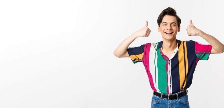 Young cheerful man smiling, showing thumbs up in approval, praise something good, standing over white background