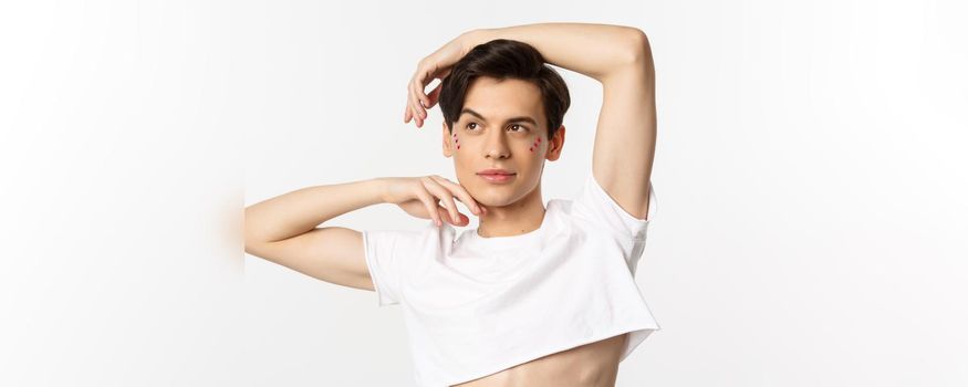 Close-up of beautiful androgynous man in crop top posing for camera, standing against white background