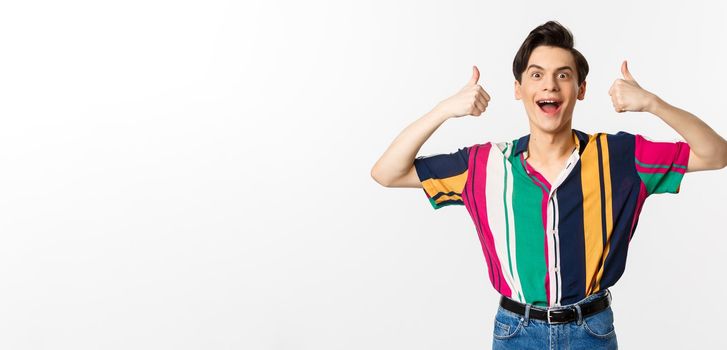Excited and amazed young man showing thumbs up with impressed expression, standing over white background