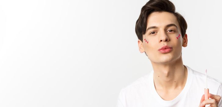 Close-up of beautiful gay man with glitter on face pucker lips after applying lip gloss, standing over white background