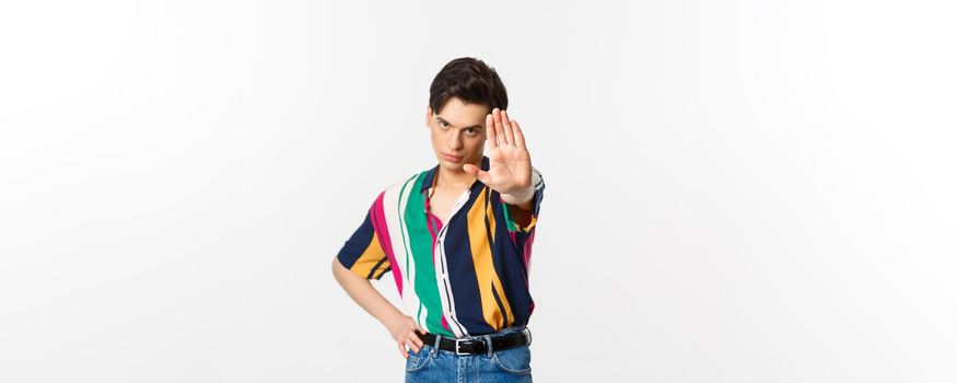 Serious young man extend hand, showing stop gesture, disapprove and prohibit action, standing over white background