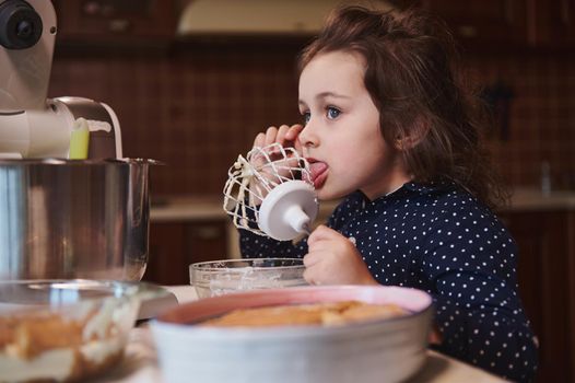 Cute little girl licks whisk with leftovers of whipped cheese cream