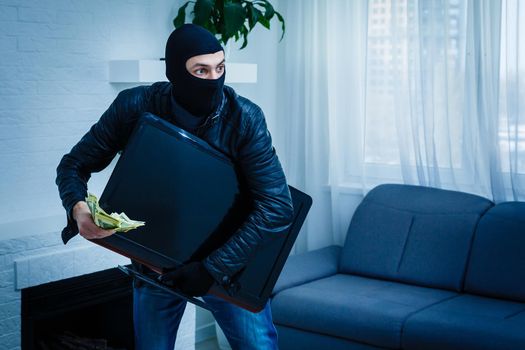 Robber man dressed in black hoodie stands with disguised face and holds a lot of money in his hands, stole a large amount, a thief man stole a TV