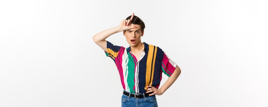 Sassy and arrogant guy showing loser sign on forehead, mocking someone, standing over white background