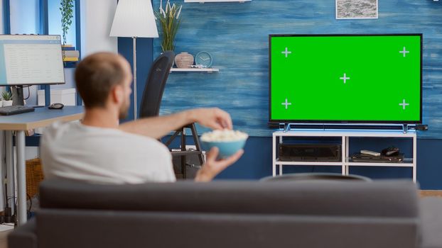 Man relaxing on couch looking at green screen on tv watching movie while eating popcorn