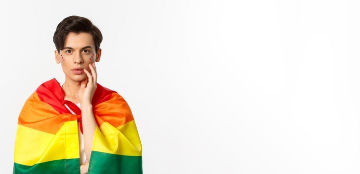 Beautiful young gay man wearing lgbt pride flag and glitter on face, touching cheek and gazing sensual at camera, white background