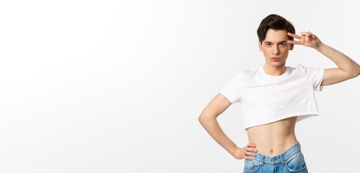 Beautiful androgynous man in crop top showing peace sign and smiling, standing over white background