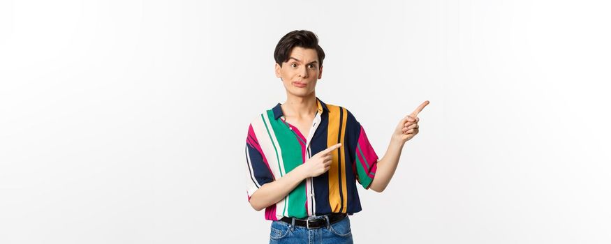 Skeptical gay man grimacing disappointed, pointing fingers right at bad product, standing over white background