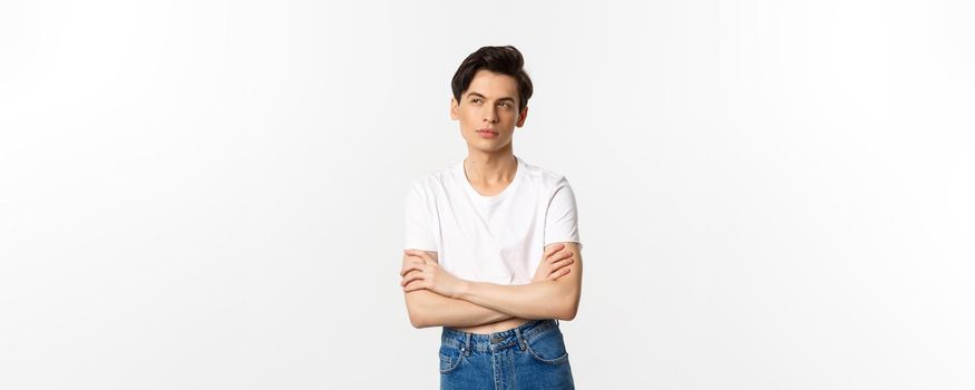 Thoughtful gay man in crop top looking at upper left corner, thinking with arms crossed on chest, standing against white background