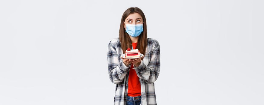 Coronavirus outbreak, lifestyle during social distancing and holidays celebration concept. Dreamy cute girl in medical mask, dreamy look up, imaging making wish as holding birthday cake with candle
