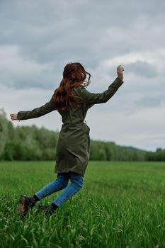 happy woman in a long raincoat jumping in a green field in cloudy weather