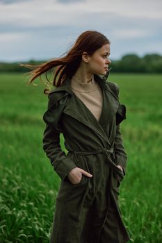 a beautiful red-haired woman stands in a field in a long dark raincoat, holding her hands in her pockets