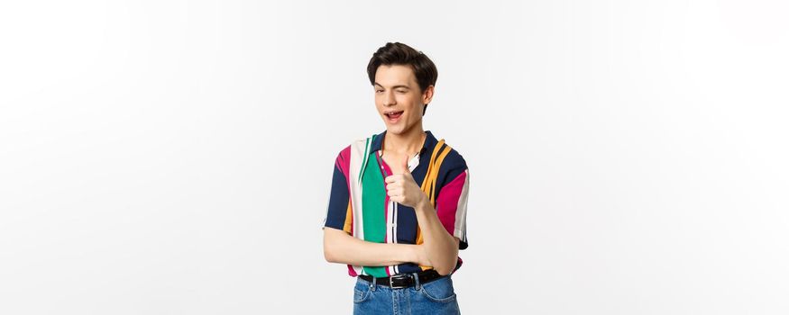 Handsome young gay man showing thumb-up and winking, encourage you, praising nice choice, standing over white background