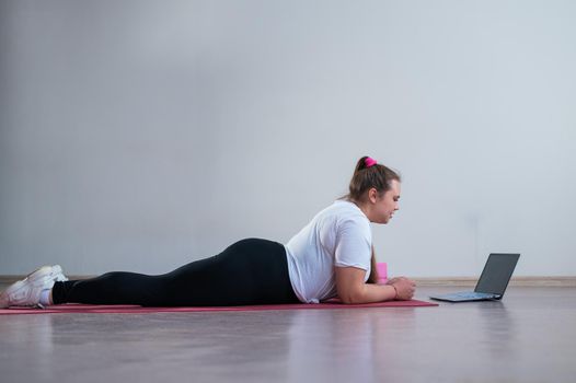 Young Plus Size Woman Stretching At Home Online. Flexible girl practices yoga and watches an online course on a laptop
