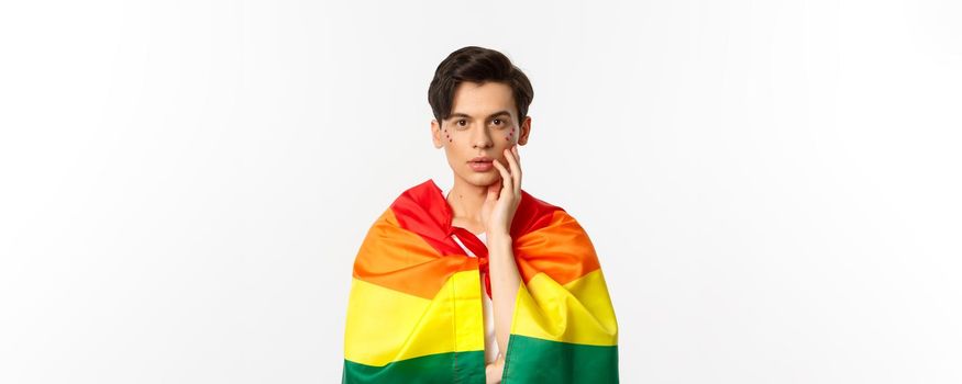Beautiful young gay man wearing lgbt pride flag and glitter on face, touching cheek and gazing sensual at camera, white background