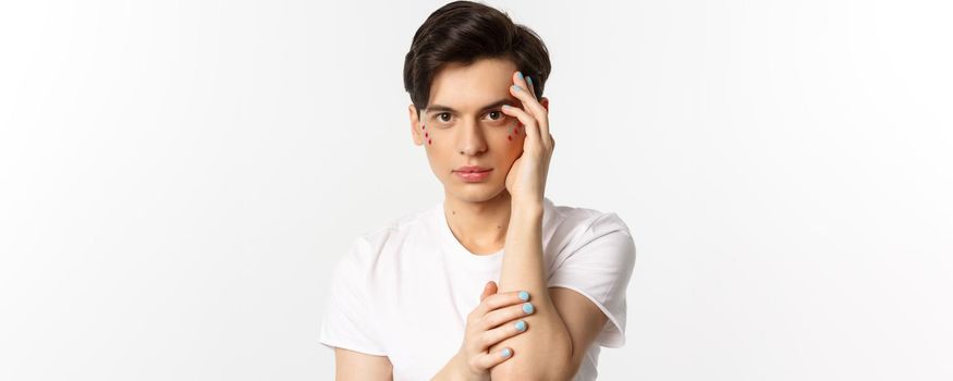 People, lgbtq and beauty concept. Close-up of beautiful queer man touching face with fingers with blue nail polish, standing over white background