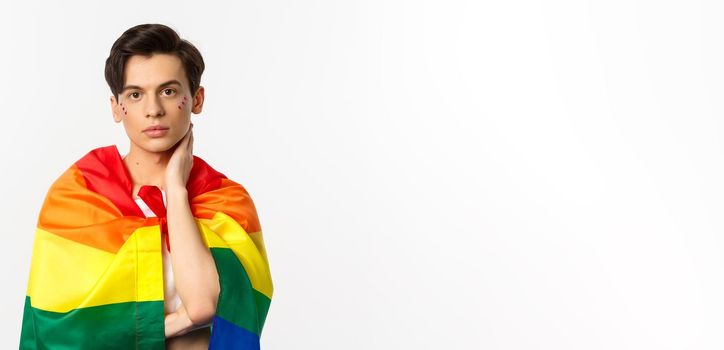 Waist-up shot of beautiful gay man with glitter on face, wrap himself with rainbow lgbt flag and touching face gently, looking at camera, white background