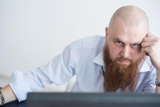 A focused bald man with a red beard stares intently at the camera. Male manager in a white shirt is angry at work.