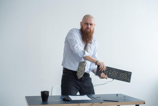 Angry bald man with a red beard in the office in a business suit crashes a computer. Manager with a nervous breakdown breaks the keyboard on the monitor.