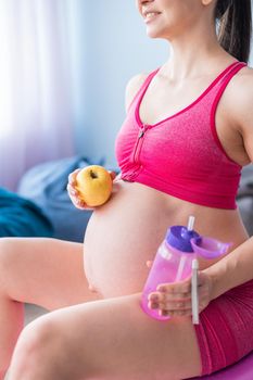 Faceless pregnant woman eating an apple after yoga. Expectant mother bites fruit and drinks water after Pilates. Girl sitting on a fitness ball.