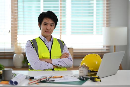 Handsome civil engineer in white shirt and yellow vest sitting with arms crossed and looking at camera.