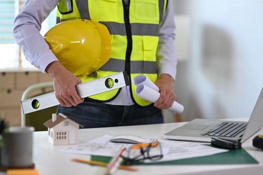 Cropped image of civil engineer holding safety helmet and blueprints standing in office.