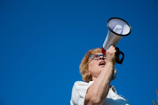 Emotional senior woman makes a speech in a megaphone on the outside. A pensioner yells into a sound amplifier on a blue background. female leader of the rally voices the conditions in the loudspeaker.