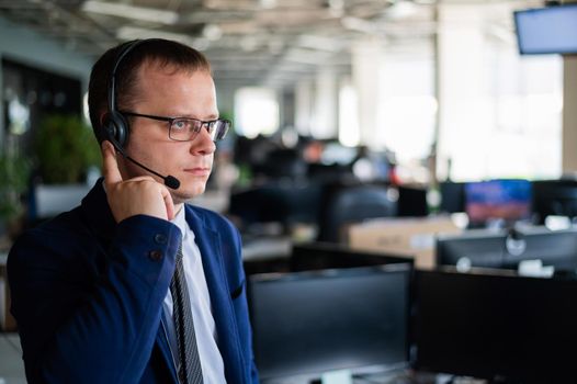 Portrait of a male call center operator in headset at workplace. A man works in an open space office answering customer calls. Support service or hotline. Agent of a telemarketing company.