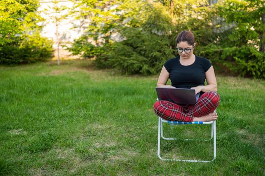 A woman with glasses sits on a folding chair and types on a laptop. Female freelancer works remotely in the garden of the cottage. Work in the open air during quarantine.