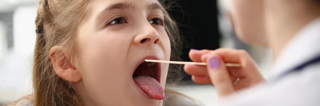 Little girl having her throat examined with special spoon at woman doctor in hospital