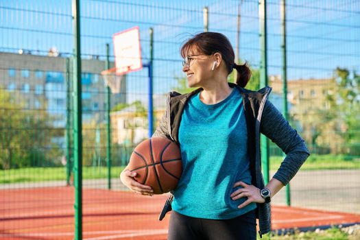 Middle aged smiling active woman with ball, near outdoor basketball court