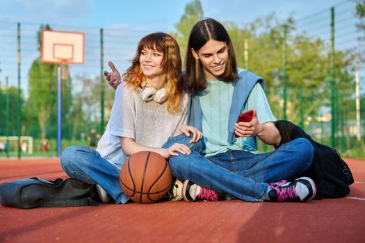 Student friends sitting on outdoor basketball court looking in smartphone