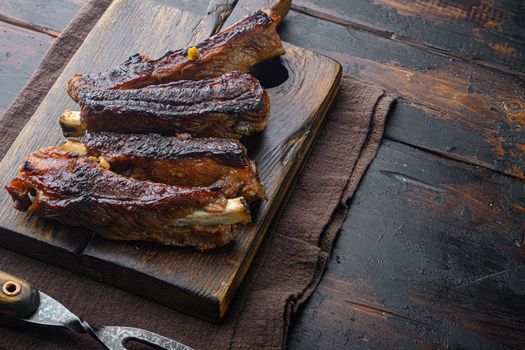 Grilled barbecue pork ribs, on wooden serving board, with barbeque knife and meat fork, on old dark wooden table background