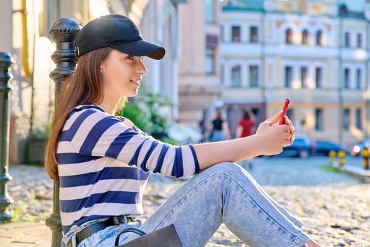 Urban style, fashionable teenage female sitting on the sidewalk using smartphone