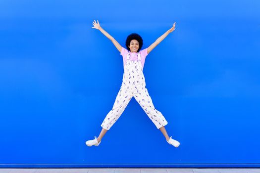 African American female jumping with raised arms against a blue urban wall.