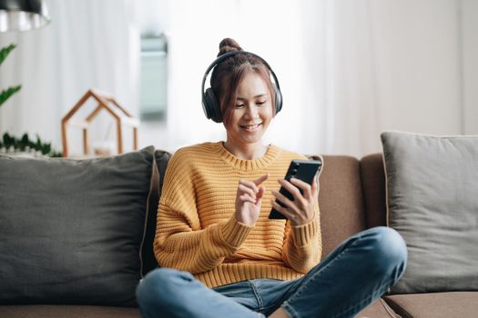Freelance asian woman with mobile phone listening music in headphones and relax at home. Happy girl sitting on couch in living room.