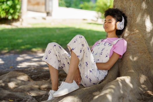 Black woman sitting in the shade of a tree relaxing and listening to music with headphones.