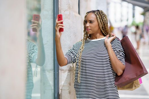 Young black female photographing a store window in a shopping street with her smartphone.