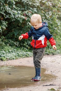 A timid little boy from the city in a red jacket, carefully trying to go the puddle.